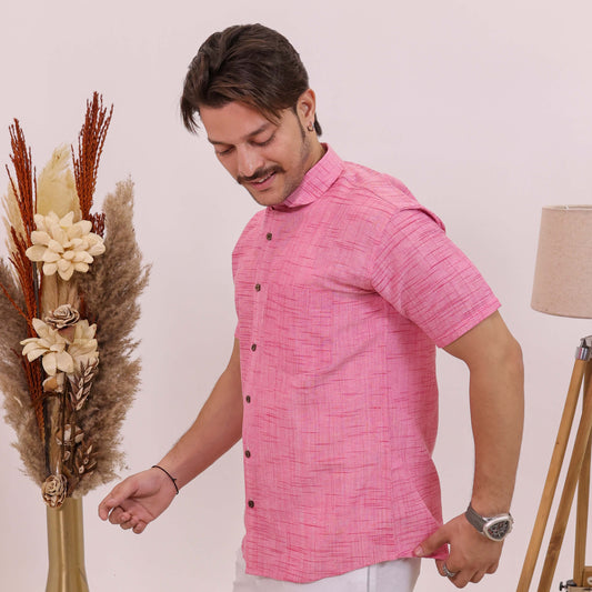 A man wearing a short-sleeve pink textured button-up shirt and white trousers, standing next to a decorative dried flower arrangement in a gold vase.