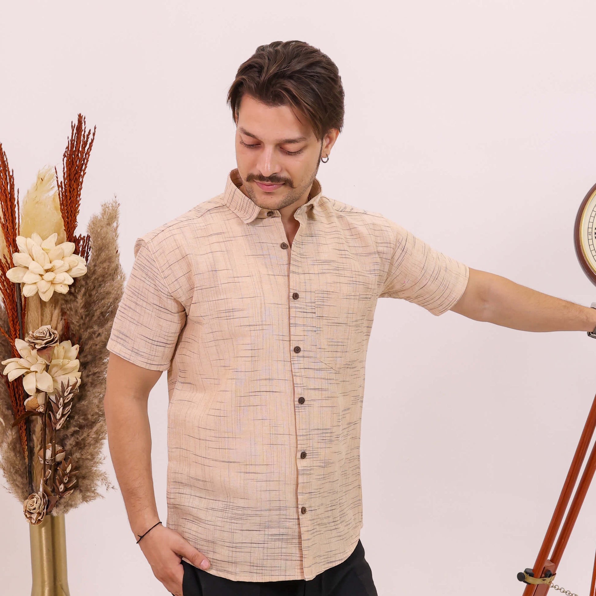 A man wearing a beige short-sleeve button-up shirt with a subtle textured pattern and black trousers, standing next to a decorative wooden tripod and a vase of dried grasses against a plain light background.