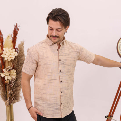 A man wearing a beige short-sleeve button-up shirt with a subtle textured pattern and black trousers, standing next to a decorative wooden tripod and a vase of dried grasses against a plain light background.